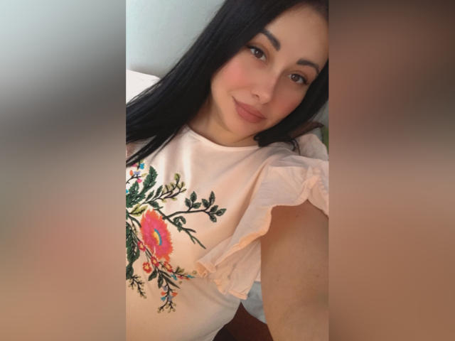 DaisySweet69 mit langen, schwarzen Haaren, heller Haut und braunen Augen, trägt ein weißes, geblümtes Hemd, lächelt in die Kamera und wirkt süß, entspannt und attraktiv..