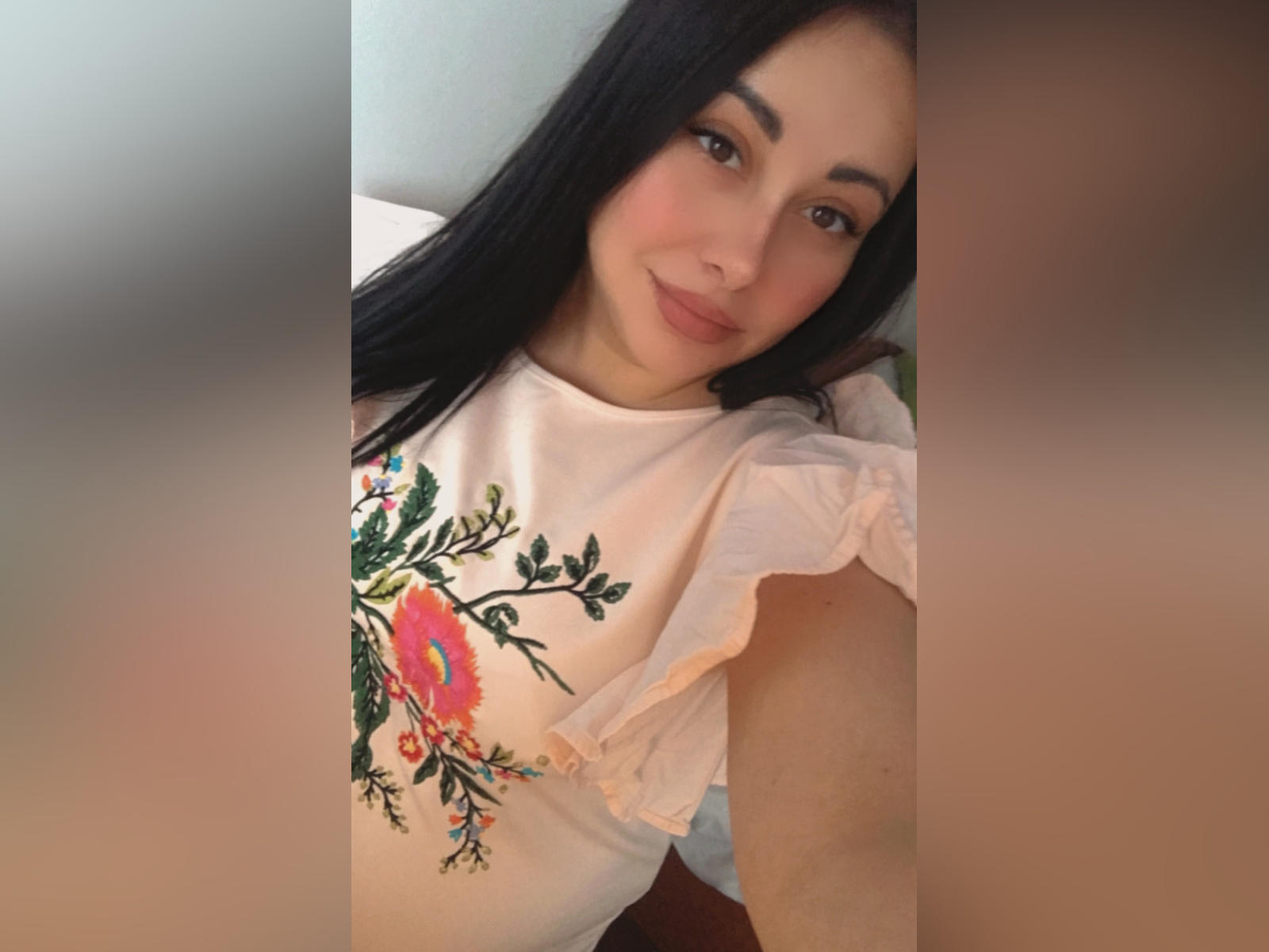 DaisySweet69, con lunghi capelli neri, pelle chiara e occhi marroni, indossa una camicia bianca con fiori, sorride alla fotocamera, mostrando un aspetto carino, rilassato e attraente..
