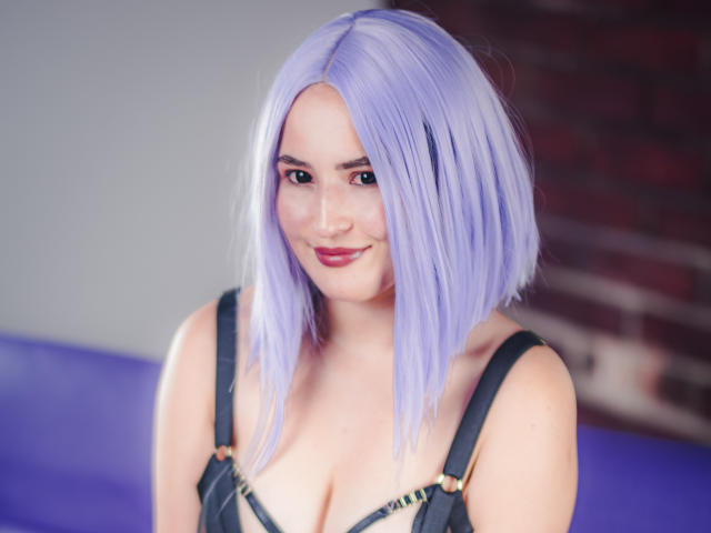 DanielaJimenes met een bleekpaars kort kapsel, lachend, draagt een zwarte jurk met bandjes, waarbij haar decolleté zichtbaar is, haar borsten zijn licht zichtbaar, roze lippenstift, bleke huid, op een zacht verlichte achtergrond van paars en baksteen..