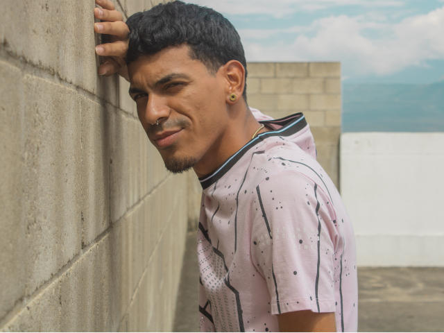 Marco Carter, avec ses cheveux noirs courts, ses boucles d'oreilles dorées, son anneau de nez et sa chemise rose à pois, est appuyé contre un mur de béton et sourit légèrement. L'arrière-plan montre un ciel nuageux..