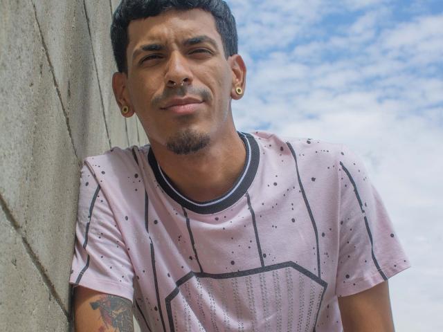 Marco Carter, com cabelo preto curto, pequena barba, piercing no nariz e brincos de ouro, vestindo uma camisa rosa com bolinhas, apoiado em uma parede de concreto, sorrindo levemente. Tatuagem no braço direito..