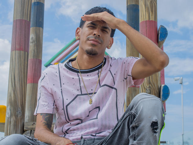 Marco Carter, con i capelli neri corti, la pelle color nocciola e un piccolo pizzetto, indossa una camicia rosa a pois, una collana d'oro e jeans strappati, stringe gli occhi e si porta una mano sulla fronte..