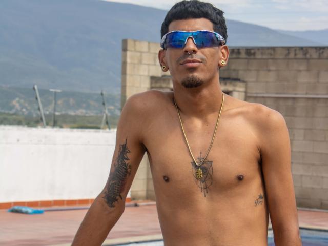 MarcoCarter, shirtless, wearing blue reflective sunglasses, gold hoop earrings, necklace with pendant, tattoos on right arm and chest, standing on rooftop, mountains in background.