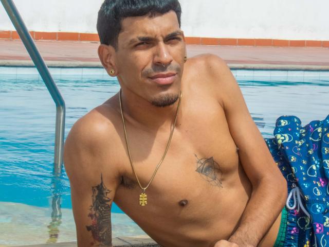 Marco Carter, torse nu, porte un collier en or, a des tatouages sur la poitrine et le bras, et une petite moustache. Il est appuyé près de la piscine, vêtu d'un maillot de bain vert, et fait une grimace..