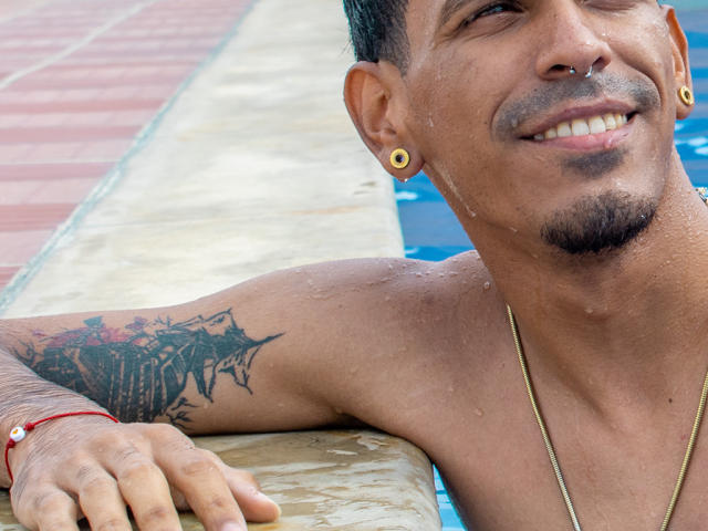 MarcoCarter, sem camisa, com cabelo curto e escuro, brincos de ouro, piercing no nariz, tatuagem no braço direito, gotas de água, sorrindo, apoiado na borda da piscina, colar de ouro, pulseira vermelha..