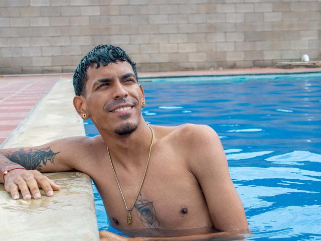 MarcoCarter, shirtless, with black short hair, small tattoos, and a gold necklace, is in a blue pool, smiling, leaning on the edge, with water droplets on his skin.