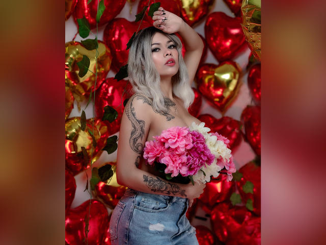 DariaDawson mit rosa und weißen Blumen, die sie vor ihrer Brust hält, trägt zerrissene Jeans, hat blondes Haar und Tattoos, im Hintergrund ein rot-goldenes Herz, ein Arm ist erhoben, sie hat einen verführerischen Ausdruck..
