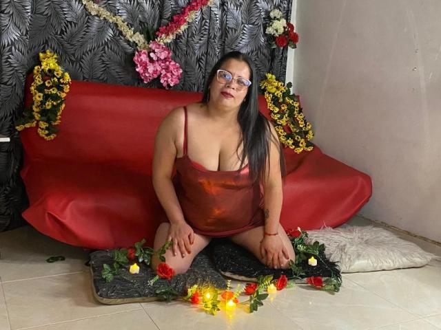 LatinCGirls with large tits, black hair, and glasses, kneeling on a red couch, wearing a shiny red dress, surrounded by flowers and candles on a tiled floor.