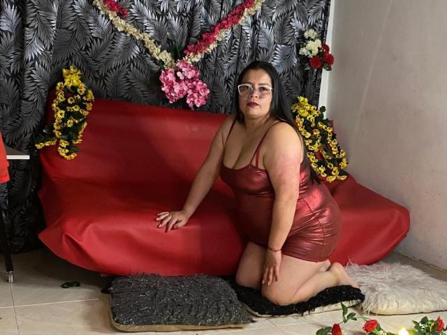 LatinCGirls a de grandes seins, des fesses rebondies, et est agenouillée sur un canapé rouge, vêtue d'une robe rouge, brillante et moulante, avec des fleurs et des coussins autour d'elle..
