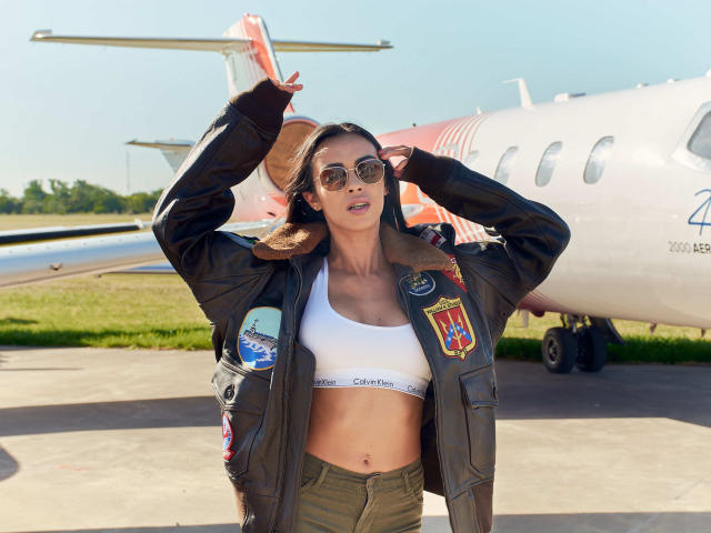 ValentinaRousse with round sunglasses, wearing a brown leather jacket with patches, a white Calvin Klein sports bra, and green pants, standing in front of a small airplane.