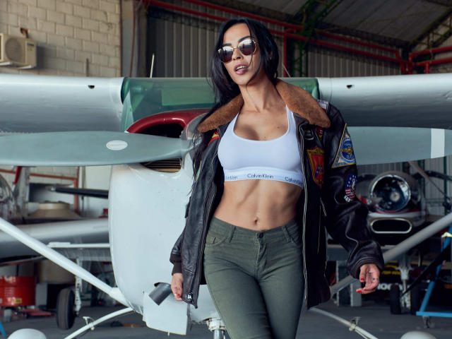 ValentinaRousse met donker haar, zonnebril, een witte sportbeha, groene broek en een zwart leren jack met bontkraag, staat voor een klein vliegtuig en laat haar getoonde buikspieren en borsten zien..