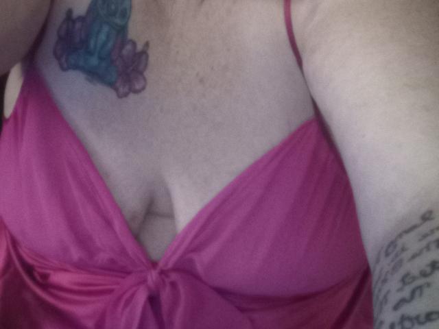 CrowButterfly's tits in a pink, tied-up top, showing a blue and purple tattoo on her right chest, with a tattooed arm and a cleavage.