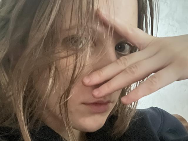 AlexandraMeow met nat, rommelig blond haar dat één oog bedekt, die haar gezicht aanraakt met een hand, gekleed in een donker shirt, met een lichtjes fronsende, serieuze uitdrukking..