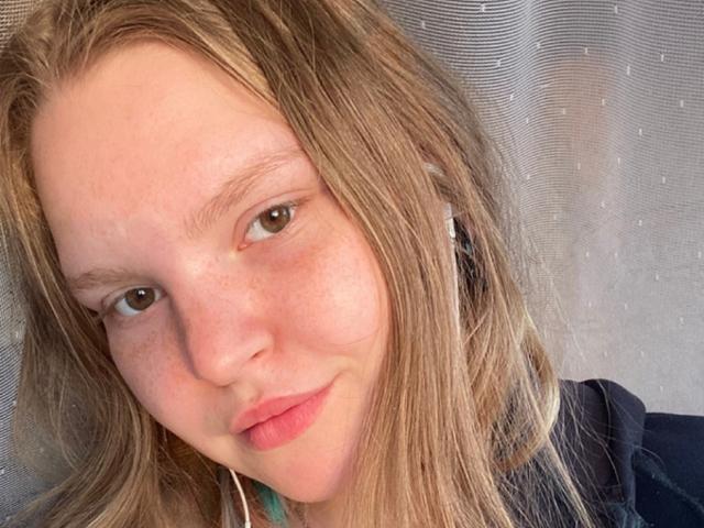 CatrinaTays Gesicht zeigt hellbraune Augen, rosafarbene Lippen und Sommersprossen, mit blonden Haaren, weißen Kopfhörern und einem leichten Lächeln. Sie trägt ein dunkles Hemd und sieht niedlich aus..