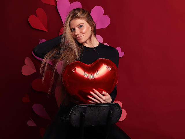 GretyArdent with long blonde hair, black long-sleeve shirt, holding shiny red heart-shaped booby balloon, sitting on black chair, smiling, red background with pink hearts.