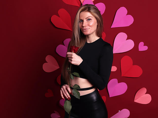 GretyArdent, avec de longs cheveux bruns, tenant une rose rouge, portant un crop top noir à manches longues et une jupe en cuir noir, debout devant un mur rouge avec des cœurs roses..