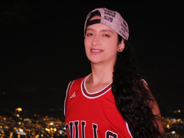 SwitchAmaraX hat langes, lockiges, schwarzes Haar, trägt ein rotes Basketballtrikot und eine weiße Kappe mit schwarzem Text, die sie verkehrt herum trägt, und lächelt leicht in die Kamera, wobei ihr Kopf leicht geneigt ist..