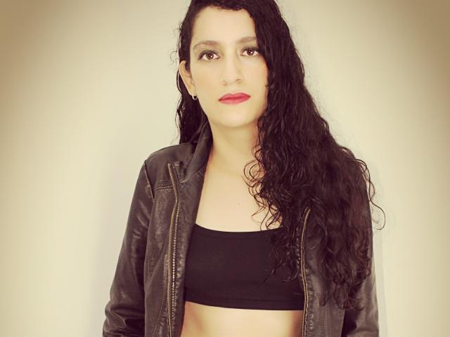 SwitchAmaraX mit langem, gewellten, schwarzem Haar, die ein schwarzes Crop-Top und eine Lederjacke trägt, steht vor einem schlichten Hintergrund, trägt roten Lippenstift und hat einen ernsten Gesichtsausdruck..