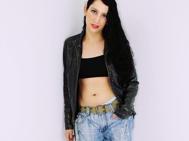 SwitchAmaraX hat langes, schwarzes Haar, trägt ein schwarzes Crop-Top, eine schwarze Lederjacke und blaue Jeans mit einem braunen Gürtel, die Hände in den Taschen, und blickt mit roten Lippen in die Kamera..