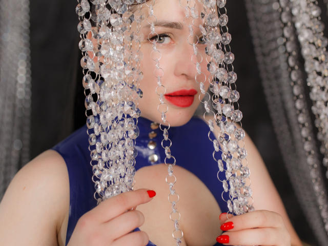 BellaSmokeyEye con lápiz labial rojo, vestido azul y tocado con cuentas de cristal, sosteniendo cuentas sobre sus senos, uñas rojas, mirando a la cámara, piel clara, fondo negro..