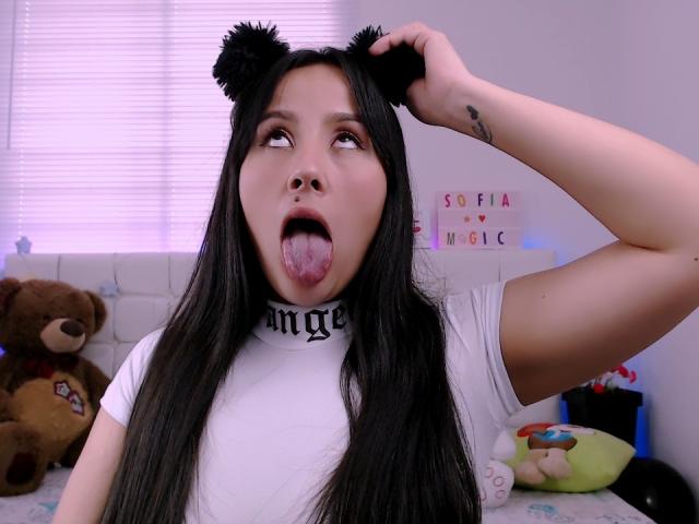 CameronGrey has long black hair, black bear ears, white shirt, and is sticking out her tongue with her tongue out, wearing a "Domme" choker. Her right arm is raised.
