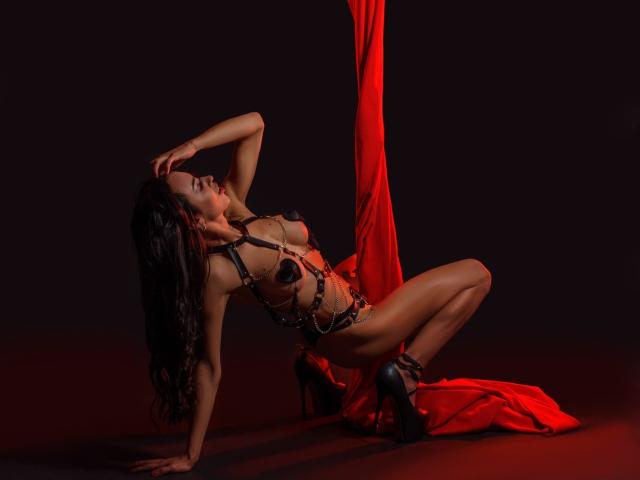 ValentinaRousse, avec ses cheveux foncés, portant de la lingerie en chaîne noire et des talons hauts, agenouillée sur de la soie rouge, les mains au sol, le dos arqué, les yeux fermés, mettant en valeur ses seins et ses fesses..
