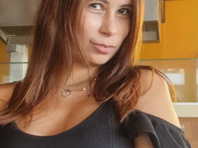 Giovanna, con cabello castaño, una blusa negra con un hombro descubierto, un collar con forma de corazón y una sonrisa sutil, mostrando sus senos en un ambiente con luz cálida..