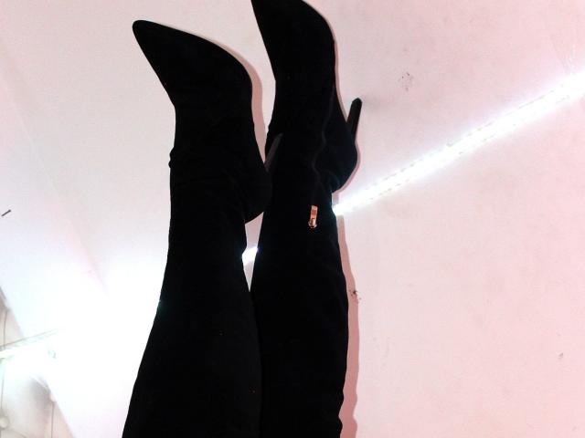 Die schwarzen, spitz zulaufenden Stiefel mit hohen Absätzen und goldenen Reißverschlüssen von SwitchAmaraX stehen vor einer weißen Wand mit einem schwachen Streifen von LED-Leuchten. Die Stiefel sind leicht nach oben geneigt..