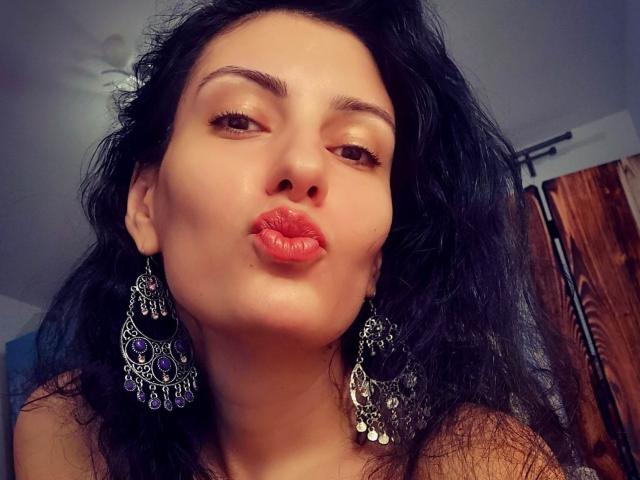 AddictedToUTS has dark wavy hair, brown eyes, and is pouting with red lipstick. She's wearing large, ornate black earrings. Her skin is light, and she looks confident.