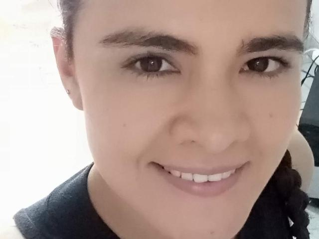 O rosto de PatyHottLove, com pele marrom clara, olhos castanhos, sobrancelhas escuras e um pequeno sorriso, vestindo uma camisa preta, mostrando uma bunda fofa, ligeiramente cheinha, ao fundo..