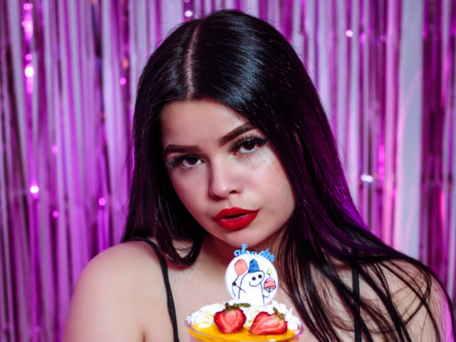 BrithneyLee with long black hair, red lipstick, and black spaghetti straps, holding a cake with strawberries and a mouse topper, looking seductively at the camera.