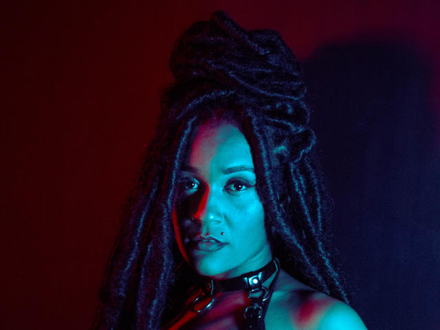 AmyStone69 met een gezicht dat blauw en rood oplicht, donkere dreadlocks, een zwarte leren choker, een neusring en een serieuze uitdrukking. Haar borsten zijn niet zichtbaar, maar haar nek en gezicht wel..