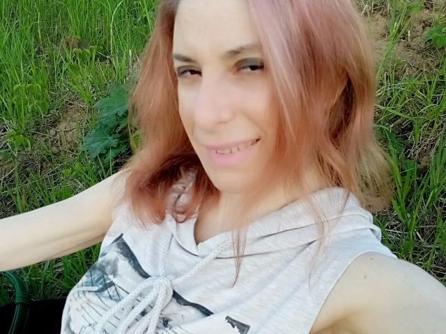 Une femme séduisante avec des cheveux roses, souriante, portant un haut blanc sans manches, montrant ses petites seins, debout dans l'herbe, prenant un selfie, son bras tendu, et un léger sourire avec un espace entre les dents..