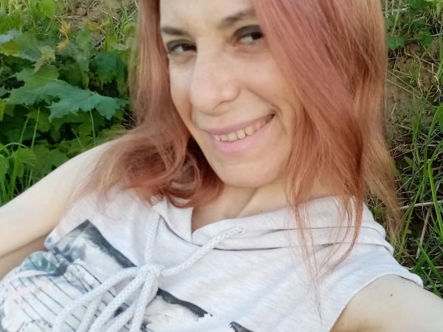 ASexyLady with pink hair, smiling, showing teeth, wearing a white hoodie with a knot, holding a selfie, green plants in background, cute, soft pink lips, light skin.