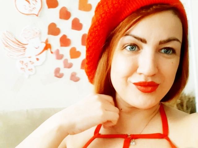 KiraXTender tiene el pelo rojo, ojos azules y lápiz labial rojo. Lleva un gorro de punto rojo y un top rojo con tirantes, mostrando sus pechos. El fondo tiene dibujos de corazones y ángeles..