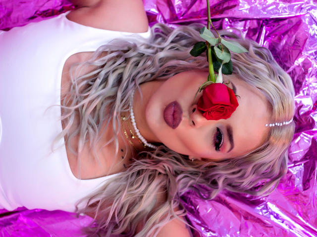 ChantalSex69, avec de longs cheveux blonds ondulés, portant un haut blanc, un collier de perles et du rouge à lèvres violet, allongée sur du plastique rose, avec une rose rouge dans la bouche..