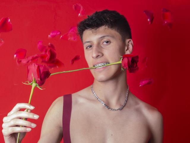 Carlos Rubiio, avec des cheveux courts et bouclés bruns, portant une chaîne en argent et une bretelle bordeaux, tenant des roses rouges dans la bouche, des pétales flottant, fond rouge, souriant, torse nu, tenant une rose dans la main gauche..