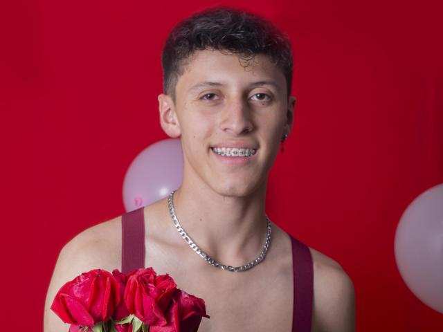 Carlos Rubiio, con capelli ricci e castani, indossa una canotta rossa, una catena d'argento, tiene in mano rose rosse, sorride mostrando l'apparecchio ortodontico, sfondo rosso, palloncini bianchi..