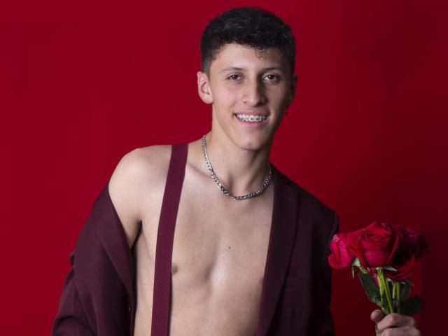 Carlos Rubiio, torse nu avec bretelles, tenant des roses rouges, souriant, avec un collier en chaîne en métal, cheveux noirs courts et bouclés, fond rouge. La poitrine de Carlos Rubiio est visible, aucun organe génital n'est montré..