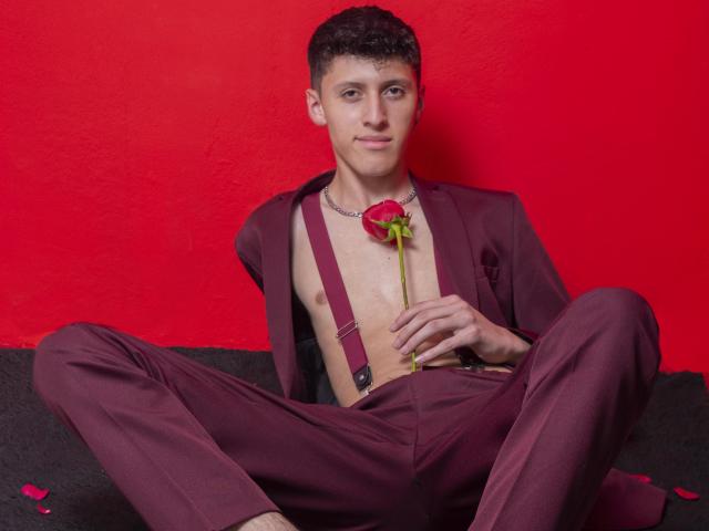 Carlos Rubiio, senza camicia, in un completo color borgogna con bretelle, che tiene in mano una rosa rossa, seduto con le gambe divaricate, di fronte a un muro rosso, indossa una collana a catena e sorride..
