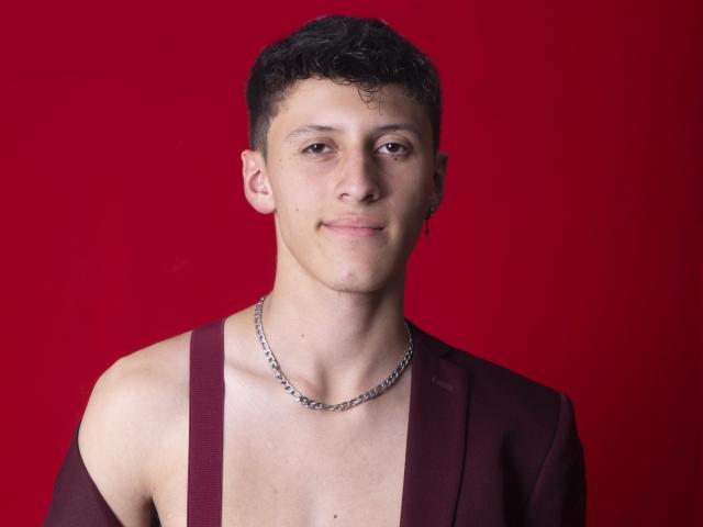 Carlos Rubiio hat lockiges, schwarzes Haar, ein leichtes Lächeln und eine Kette um den Hals. Er trägt ein tief burgunderfarbenes Oberteil mit freiliegenden Schultern vor einem einfarbig roten Hintergrund..