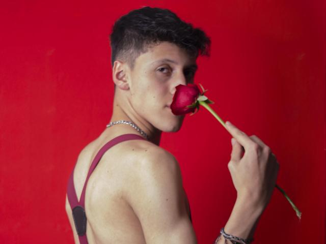 CarlosRubiio with short curly black hair, holding a red rose to his mouth, wearing a maroon strap top and silver necklace, against a solid red background.