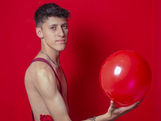 CarlosRubiio with short curly brown hair, wearing suspenders and holding a shiny red balloon, looking at the camera with a slight smile, red background.