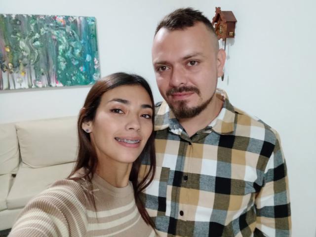HannaAndHarri, ein süßes Paar, Selfie. Hanna hat dunkelbraunes Haar, kleine Brüste, trägt ein beiges gestreiftes Oberteil und lächelt. Harri hat eine Glatze, einen Bart und trägt ein kariertes Hemd..