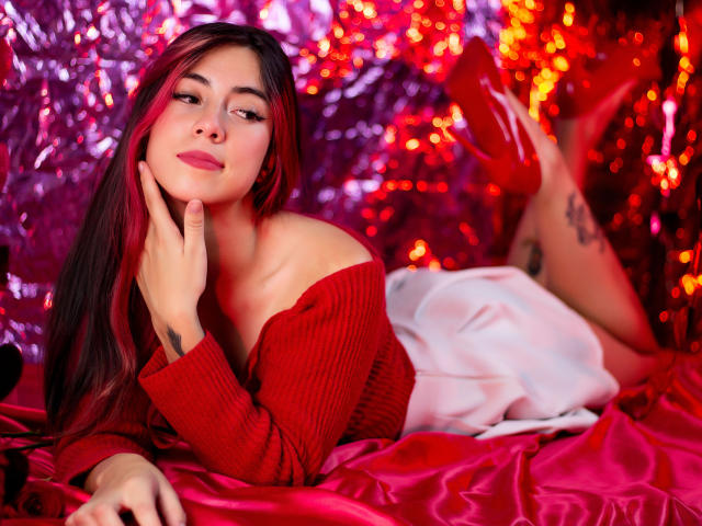JuliaGarciax with long red and black hair, red off-shoulder sweater, white skirt, red heels, laying on red satin, hand on cheek, looking at camera, tattoos visible on leg.