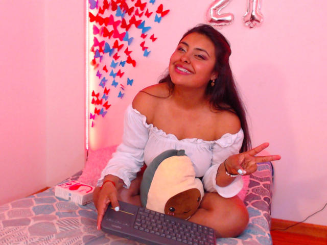 AngelaRoggers, avec des cheveux foncés, un haut blanc à épaules dénudées, souriante, tenant un clavier, un jouet en peluche, faisant le signe de la paix, dans une pièce rose, avec des décorations de papillons rouges et bleus, et un ballon avec le chiffre 2..