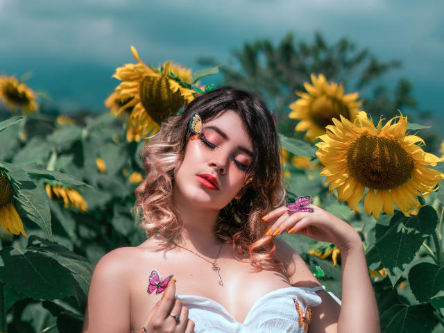 MaraMaison with wavy blonde and brown hair, wearing a white off-shoulder top, holding a butterfly, surrounded by sunflowers, with a butterfly hairpin and red lipstick.