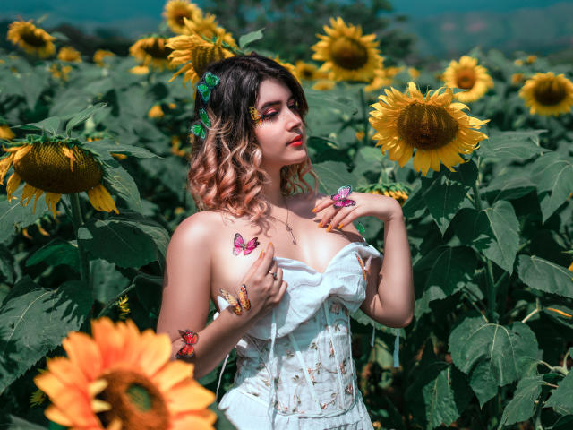 MaraMaison, com cabelos cacheados rosa e marrons, vestindo um vestido branco sem alças com estampas florais, está em um campo de girassóis, cercada por borboletas em seu peito e mãos..
