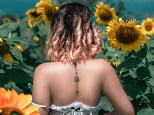 A MaraMaison está de volta, mostrando uma tatuagem na coluna, com cabelo cacheado rosa, girassóis e folhas verdes. Ela está vestindo um vestido branco com alças, e os seios estão cobertos..
