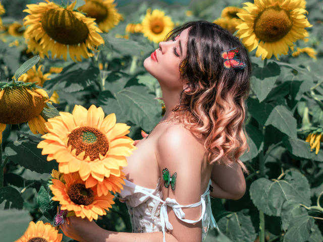 MaraMaison met golvend bruin haar, een rode vlinderhaarspeld, zonnebloemen vasthoudend, een witte jurk met een open schouder, een groene vlindertatoeage, ogen gesloten, lachend, zonnebloemen in een veld, een zonnige dag..
